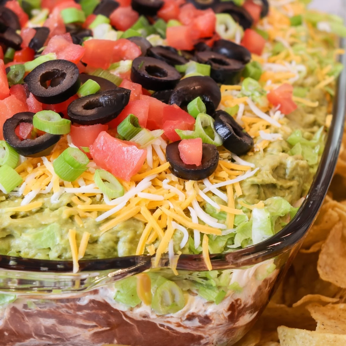 A chilled Ultimate Seven Layer Taco Dip is topped with shredded cheese, tomatoes, and sliced black olives for a festive appetizer.