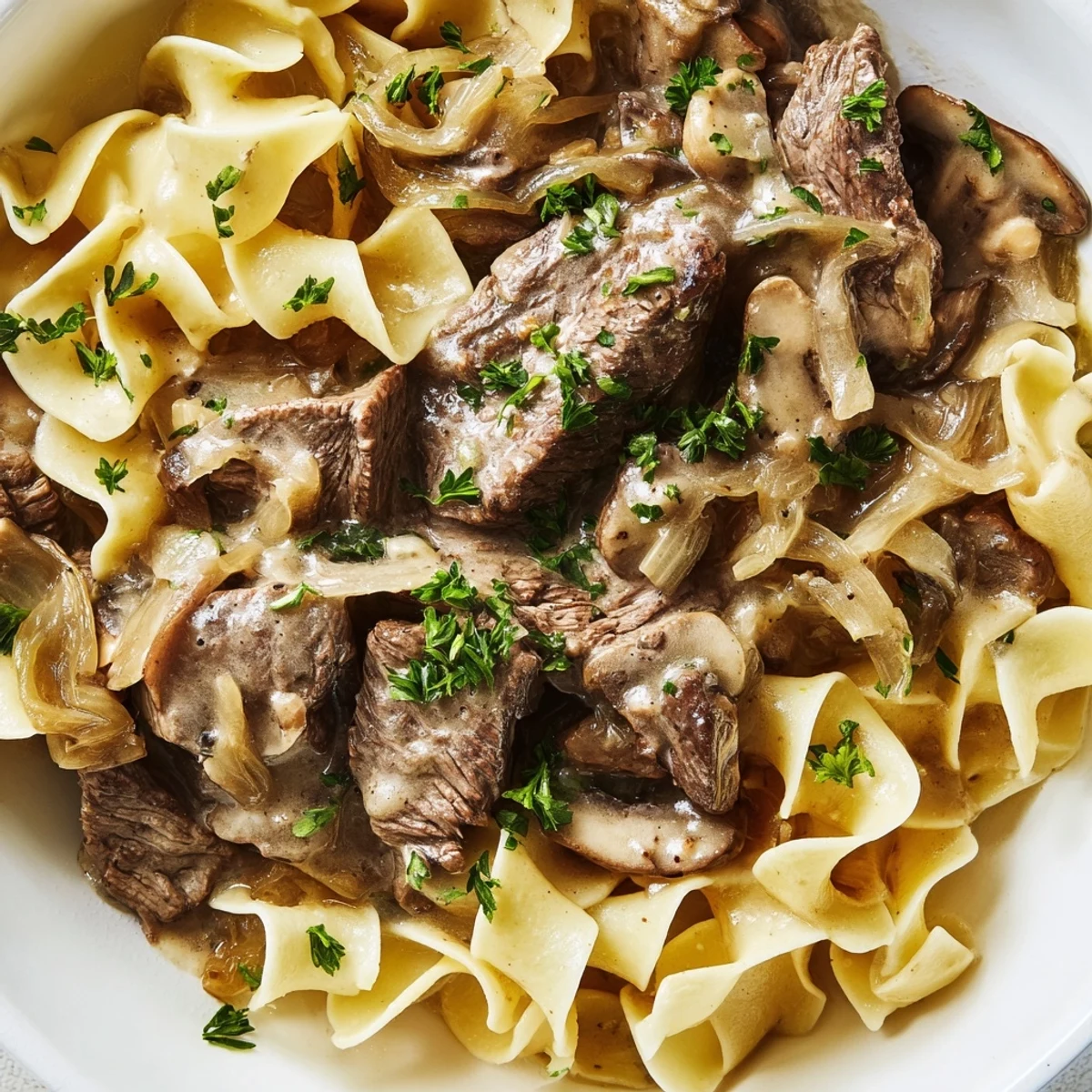 A close-up view of tender beef stroganoff with egg noodles, sautéed mushrooms, and onions covered in a savory, creamy sauce.