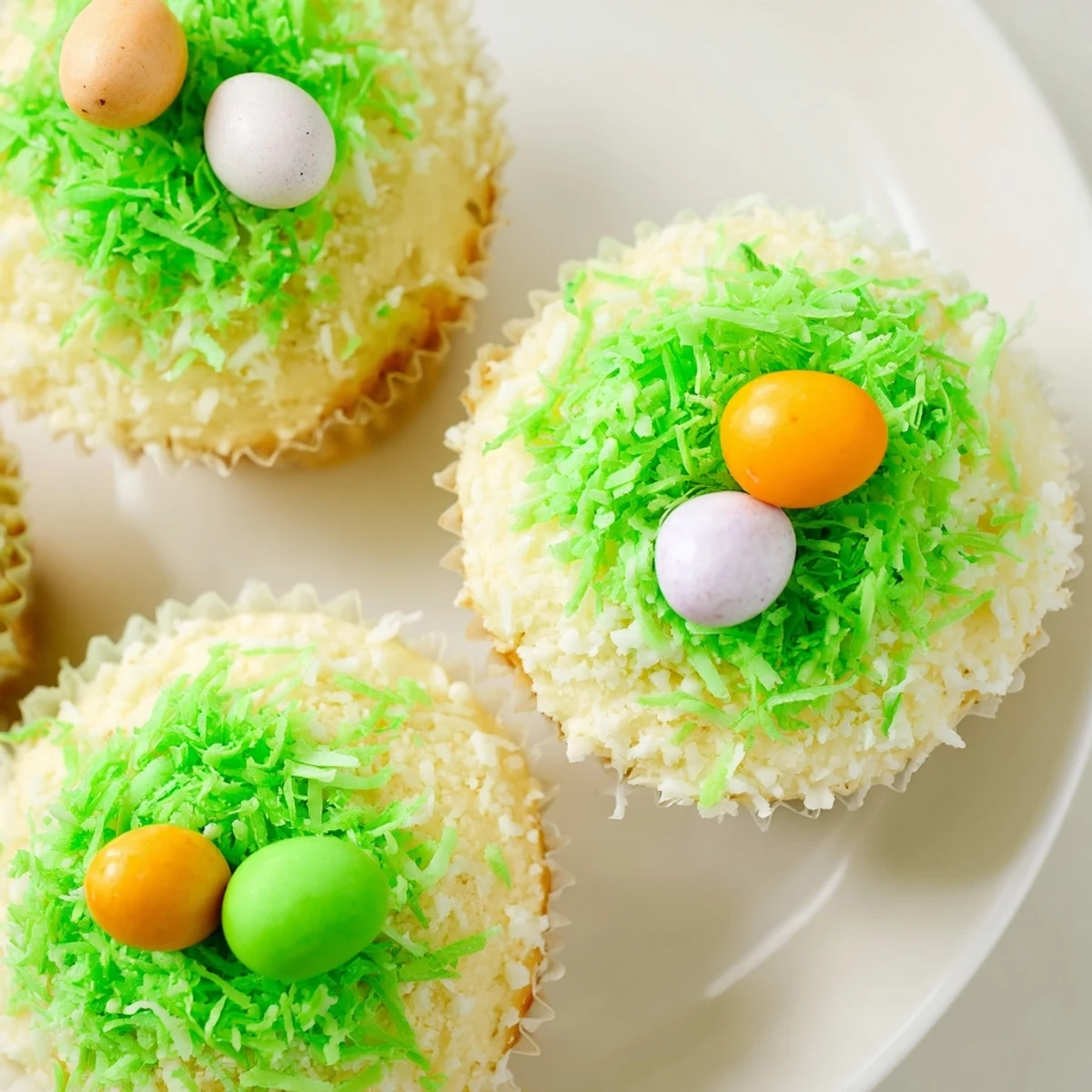 Brightly frosted Easter Basket Cupcakes topped with green-tinted coconut grass and colorful candy eggs, arranged on a pastel serving platter.