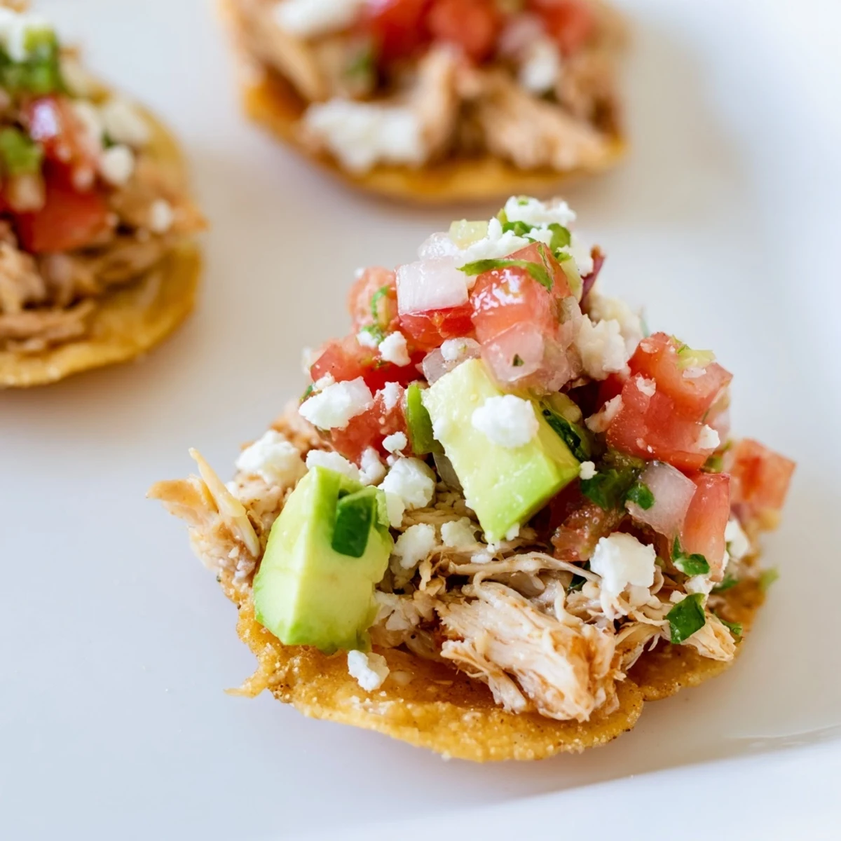 Golden-brown, crunchy mini chicken tostadas topped with fresh pico de gallo, diced avocado, and a drizzle of sour cream on a serving platter.  