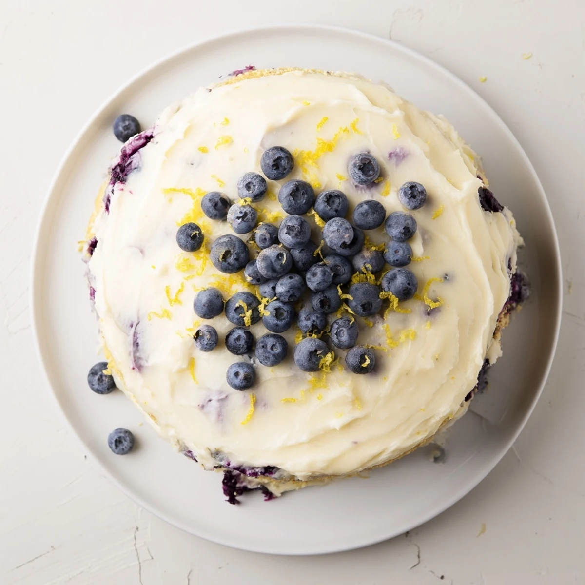 Two-layer lemon blueberry cake topped with creamy lemon frosting and scattered fresh berries on white plate