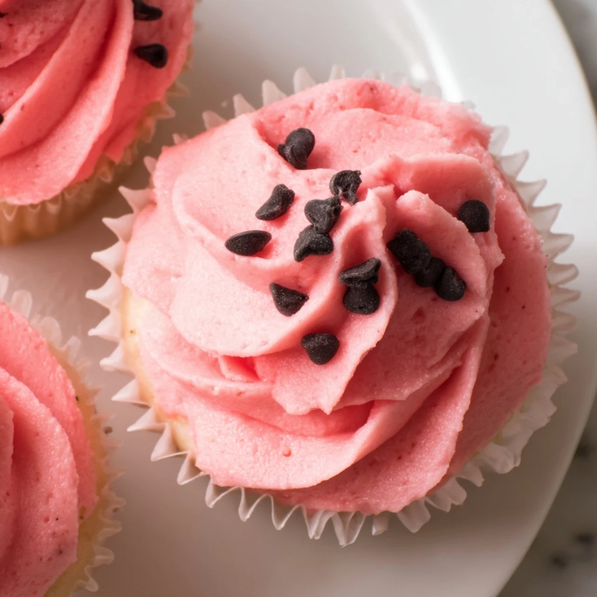 Pink watermelon cupcakes topped with creamy pink frosting and chocolate chip seeds on a white plate